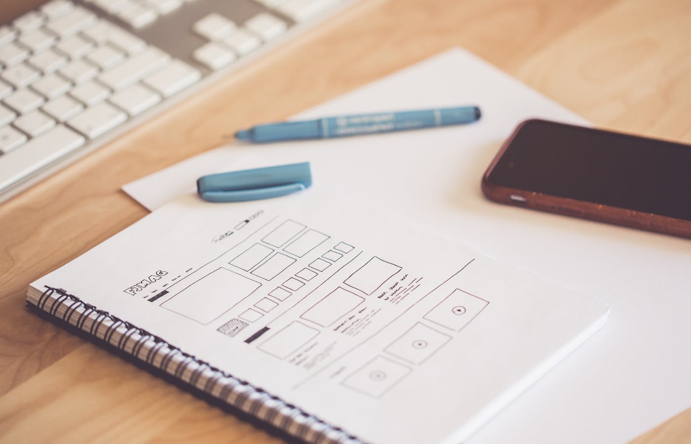 Notebook with homepage wireframe sketches beside a phone, pen, and keyboard.